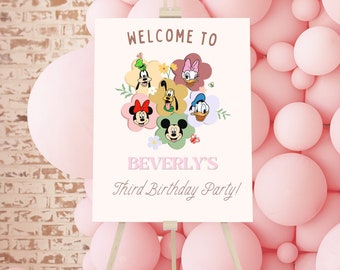 May include: Welcome to Beverly's Third Birthday Party! A white sign with a colorful illustration of Mickey Mouse and friends in a floral wreath. The sign is on a wooden easel in front of a pink balloon wall.
