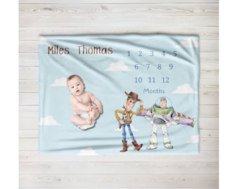Puede incluir: Una manta azul de hitos para bebés con un patrón de nubes blancas. La manta presenta a un bebé, Woody y Buzz Lightyear de Toy Story. La manta tiene el texto "Miles Thomas" y los números del 1 al 12 para hacer un seguimiento del crecimiento de un bebé.