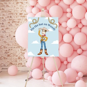May include: A blue and white game board with a cartoon image of Woody from Toy Story. The game is called "Pin the hat on Woody". The board is on an easel in front of a wall of pink balloons.