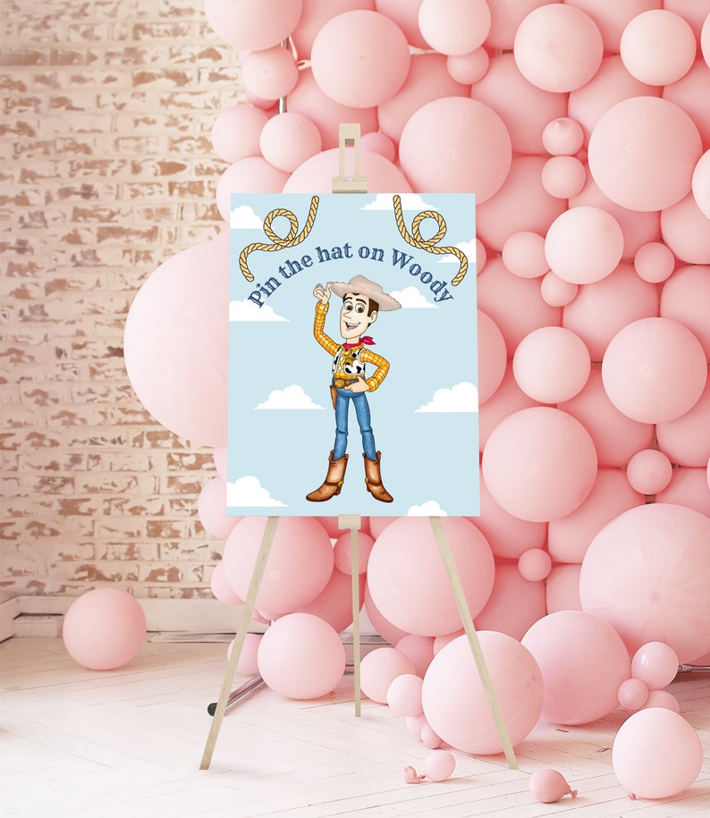 May include: A blue and white game board with a cartoon image of Woody from Toy Story. The game is called "Pin the hat on Woody". The board is on an easel in front of a wall of pink balloons.