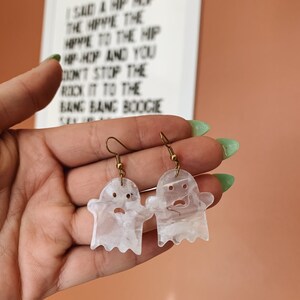 May include: A pair of white ghost-shaped earrings with a marbled effect. The earrings are made of resin and have gold-toned hooks.