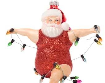 May include: A Christmas ornament depicting Santa Claus in a red sequined swimsuit, holding a string of colorful Christmas lights. Santa is wearing a red hat and has a white beard. The ornament is designed to be hung on a Christmas tree.