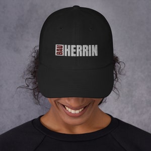 Könnte beinhalten: Schwarze Baseballkappe mit einem weißen gestickten Logo, das "BAU HERRIN" in einem roten Kasten zeigt.