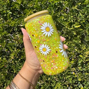 May include: A yellow glass tumbler with a wooden lid and a white daisy pattern. The tumbler is filled with yellow glitter and small white, pink, and orange glitter.