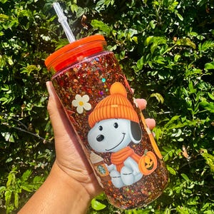 Puede incluir: Un vaso transparente lleno de purpurina marrón y naranja, con un gráfico de un perro de dibujos animados con gorro y bufanda naranja, sosteniendo un café junto a una pequeña calabaza y una flor blanca. Tiene tapa naranja y pajita transparente.