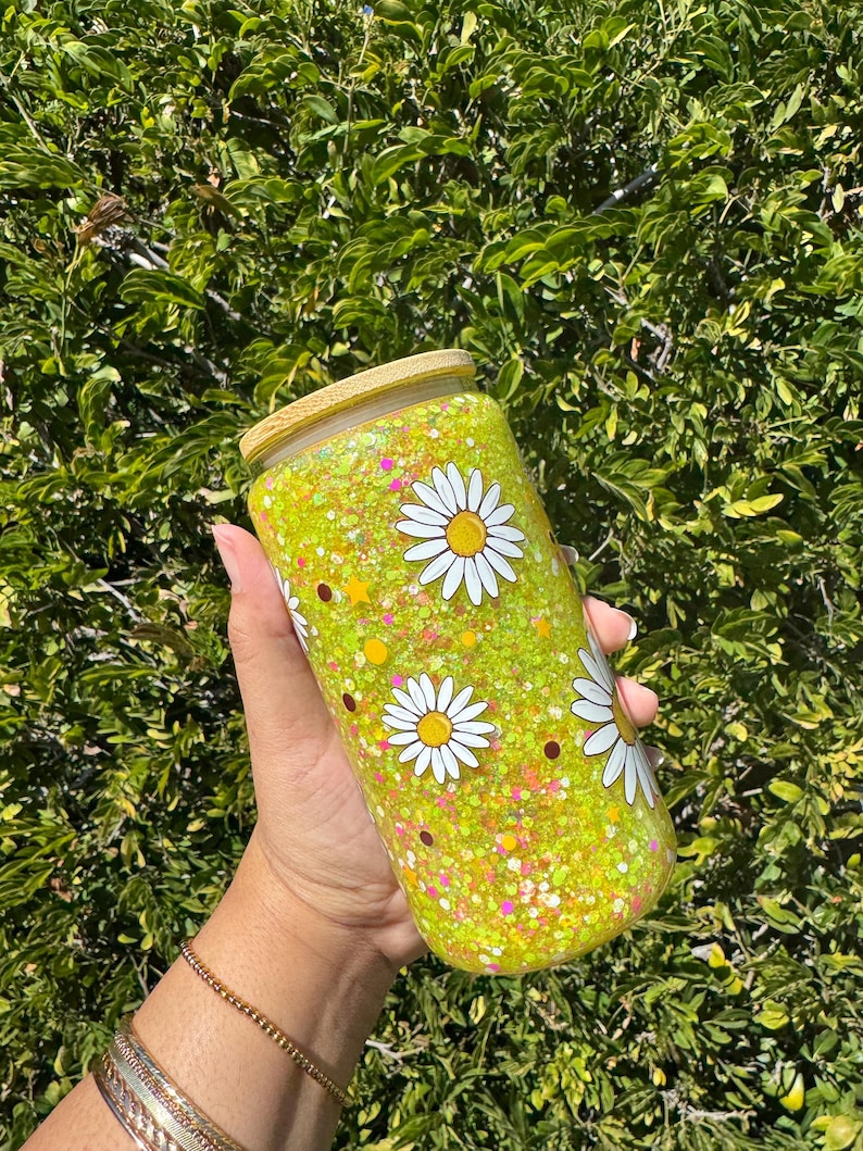 May include: A yellow glass tumbler with a wooden lid and a white daisy pattern. The tumbler is filled with yellow glitter and small white, pink, and orange glitter.