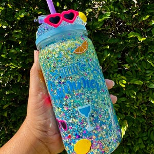 May include: A blue glitter-filled mason jar tumbler with a straw and heart-shaped sunglasses on the lid. The jar is decorated with fruit and cocktail-themed stickers and the words "Hello Summer".