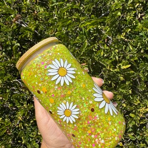 May include: A yellow glitter tumbler with white daisy flower designs and a bamboo lid. The tumbler is cylindrical and filled with glitter. The background is a green leafy bush.