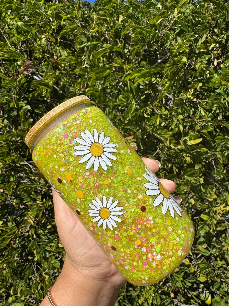 May include: A yellow glitter tumbler with white daisy flower designs and a bamboo lid. The tumbler is cylindrical and filled with glitter. The background is a green leafy bush.