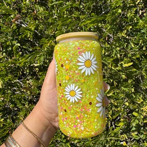 May include: A clear glass tumbler with a yellow-green glitter interior and a bamboo lid. The tumbler is decorated with white daisy flower graphics. The background is a green leafy bush.
