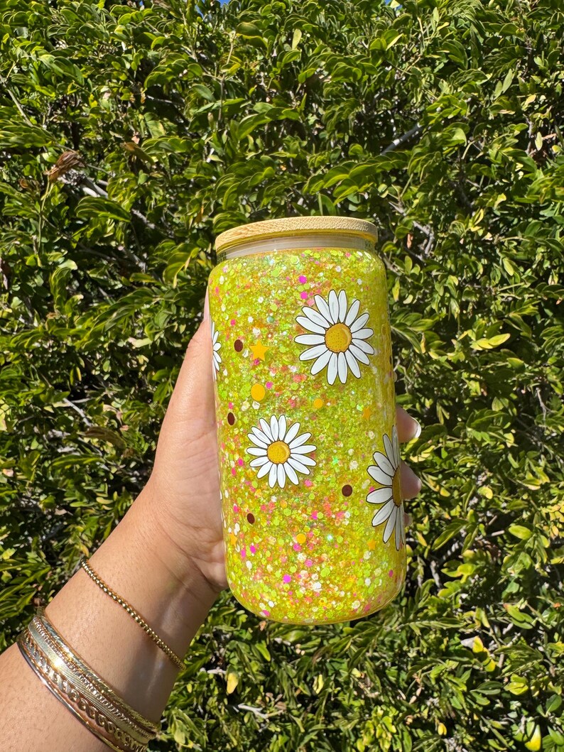 May include: A clear glass tumbler with a yellow-green glitter interior and a bamboo lid. The tumbler is decorated with white daisy flower graphics. The background is a green leafy bush.