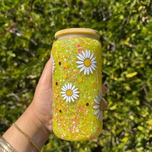 May include: A clear glass tumbler with a bamboo lid, filled with yellow glitter and daisy decals. The tumbler is decorated with white daisies with yellow centers and brown dots. The tumbler is held by a person.