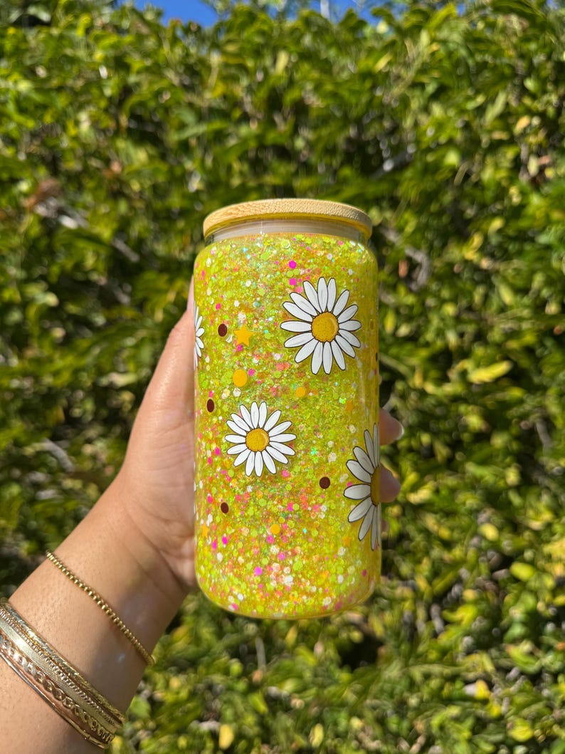May include: A clear glass tumbler with a bamboo lid, filled with yellow glitter and daisy decals. The tumbler is decorated with white daisies with yellow centers and brown dots. The tumbler is held by a person.