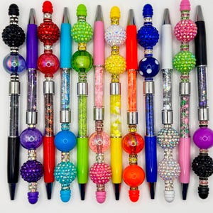 Puede incluir: Una colección de bolígrafos decorativos con varios diseños coloridos. Cada bolígrafo presenta una combinación de colores diferente, con adornos como pedrería y cuentas. Los bolígrafos tienen un barril negro elegante y están rematados con elementos decorativos.
