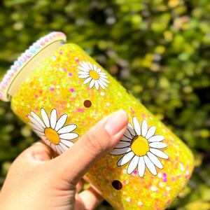 May include: A yellow glitter tumbler with white daisy flower graphics. The tumbler has a clear lid with a decorative rim. The background is blurred green foliage.