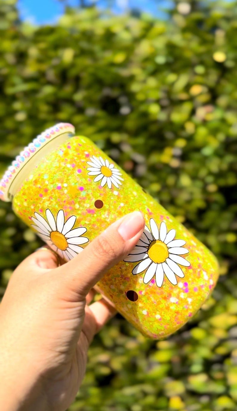 May include: A yellow glitter tumbler with white daisy flower graphics. The tumbler has a clear lid with a decorative rim. The background is blurred green foliage.