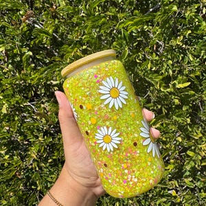 May include: A clear glass tumbler with a bamboo lid, filled with yellow glitter and daisy flower decals. The tumbler is approximately 6 inches tall and is held against a backdrop of green foliage.