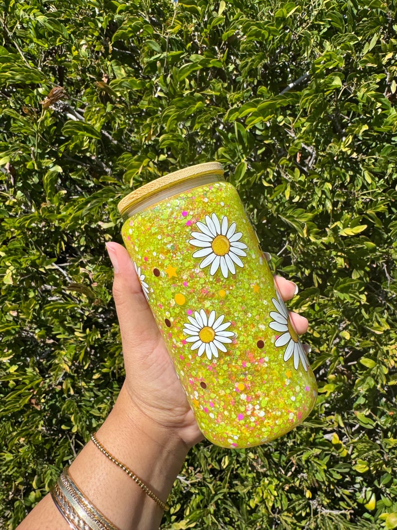 May include: A clear glass tumbler with a bamboo lid, filled with yellow glitter and daisy flower decals. The tumbler is approximately 6 inches tall and is held against a backdrop of green foliage.