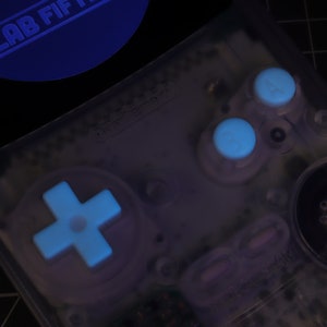 May include: A clear, translucent Game Boy Color with a white cross-shaped directional pad, two white buttons, and a speaker grill. The buttons and directional pad are glowing blue.