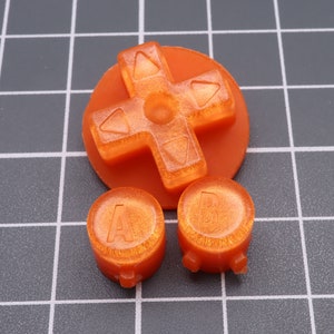 Könnte beinhalten: Orangefarbene Plastik-Videospiel-Controller-Knöpfe. Die Knöpfe sind mit A und B beschriftet. Der Richtungspad ist kreuzförmig mit vier Pfeilen.
