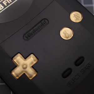May include: Close-up of a black Nintendo Game Boy with gold buttons, a cross-shaped directional pad, and the text "SELECT START" on the bottom right.