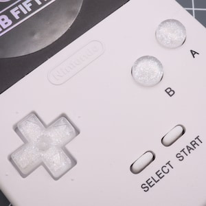 Könnte beinhalten: Nahaufnahme eines weißen Nintendo Game Boy mit einer schwarz-weißen Spielkassette. Der Game Boy hat einen weißen kreuzförmigen Steuerkreuz, zwei weiße Knöpfe mit der Aufschrift "A" und "B" und zwei weiße Knöpfe mit der Aufschrift "SELECT" und "START".