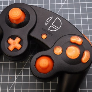 May include: A black GameCube controller with orange buttons. The controller has a white Nintendo logo and the words "START/PAUSE" printed on it.