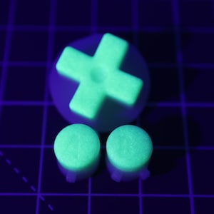 May include: Three glow-in-the-dark green buttons for a video game controller. The buttons are round and have a textured surface. One button is shaped like a plus sign.