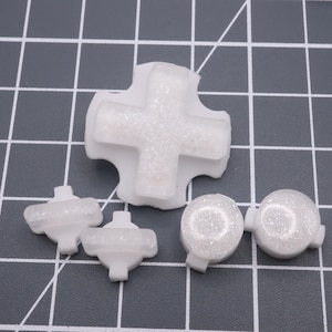 May include: Set of five white plastic buttons with a textured surface. The buttons are shaped like a cross, a circle, and a rectangle.