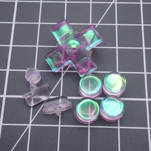 May include: A collection of iridescent replacement buttons and directional pad for a gaming controller. The buttons are round and the directional pad is cross-shaped. The colors shift between purple, green, and blue. The items are made of translucent plastic.