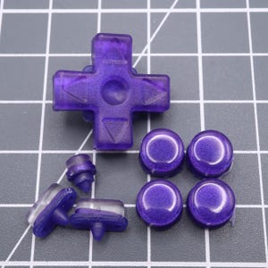 Op de afbeelding: Een set paarse knoppen voor een videogamecontroller. De set bevat een directionele pad, vier face-knoppen en twee schouderknoppen.