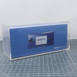 Può includere: Una teca di esposizione in acrilico trasparente con una Game Boy Micro blu all'interno. La teca è su una superficie grigia a griglia. La Game Boy Micro ha un design blu e argento con il testo "GAME BOY" sul davanti.