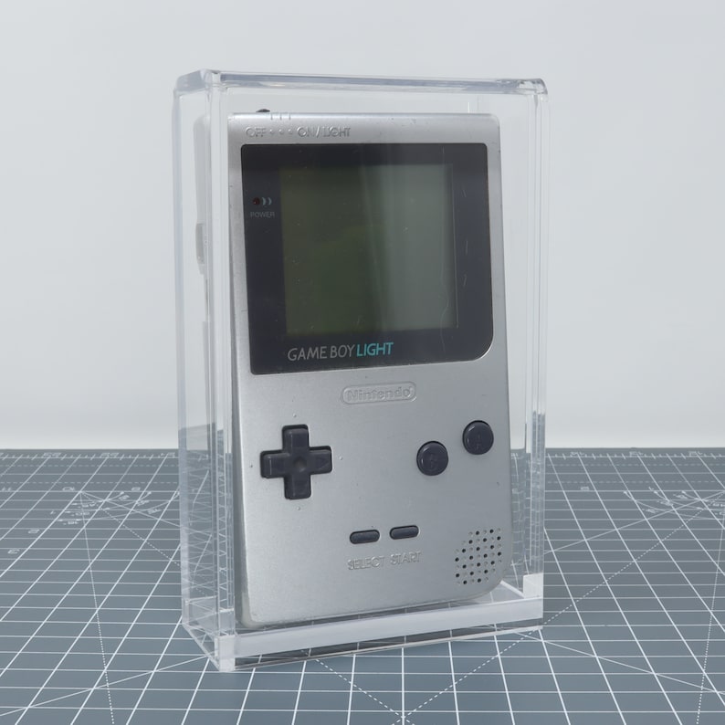 Pu&ograve; includere: Una console per videogiochi portatile Game Boy Light argentata in una teca di plastica trasparente. La console ha uno schermo bianco e nero, un pad direzionale nero e pulsanti neri. Il testo "GAME BOY LIGHT" e "Nintendo" &egrave; stampato sulla console.