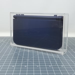 May include: A dark blue Nintendo 3DS handheld gaming console in a clear acrylic display case.