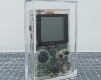 May include: A clear Game Boy Pocket video game console in a clear acrylic display case. The console has a black screen and green buttons.