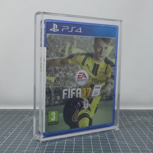 Puede incluir: Una caja de juego de PS4 para FIFA 17, exhibida en una vitrina de acrílico transparente. La portada del juego presenta a un futbolista con una camiseta amarilla. La caja tiene un borde azul y el logotipo de PS4 es visible.