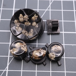 Peut inclure: Un ensemble de boutons de manette de jeu à thème crâne noir et blanc. L'ensemble comprend un grand bouton rond, quatre boutons ronds plus petits et un petit bouton rectangulaire.