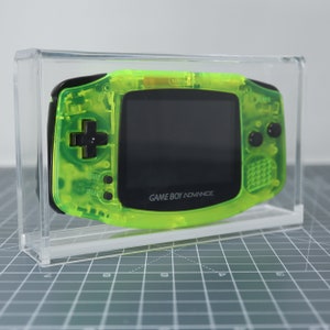 Może przedstawiać: Jasnozielona przenośna konsola do gier Game Boy Advance w przezroczystej akrylowej gablocie. Konsola ma czarny ekran i czarne przyciski. Na konsoli widnieje napis "GAME BOY ADVANCE".