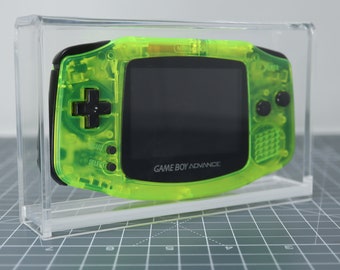 May include: A bright green Game Boy Advance handheld video game console in a clear acrylic display case. The console has a black screen and black buttons. The text "GAME BOY ADVANCE" is printed on the console.