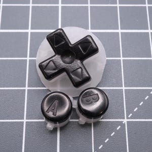 May include: A set of black replacement buttons for a video game controller. The set includes a directional pad, an 'A' button, and a 'B' button.