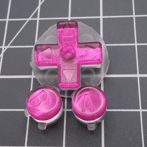 May include: A set of translucent pink game controller buttons, including a directional pad and two circular buttons marked with "A" and "B". The buttons are made of a clear, shiny material, designed for a handheld gaming device.