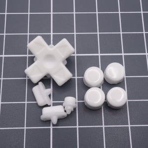 May include: A set of white plastic replacement buttons for a video game controller. The set includes a directional pad, four face buttons, and two small buttons.