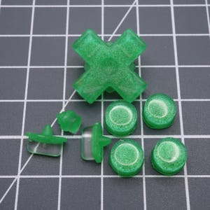 Op de afbeelding: Een set groene en transparante plastic knoppen voor een videogamecontroller. De knoppen zijn rond en hebben een getextureerd oppervlak. De knoppen zijn in verschillende maten en vormen.
