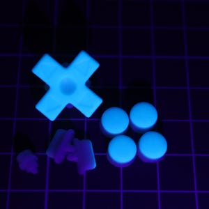 May include: A collection of glowing blue plastic components, including an X-shaped piece, four circular pieces, and other small parts. The objects are illuminated against a dark background with a grid pattern.