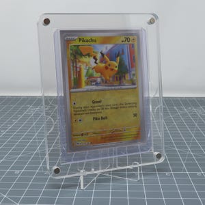 Op de afbeelding: Een transparante acryl display met een standaard, waarop een Pokemon trading card te zien is met Pikachu, een gele elektrische muis Pokemon. De kaart heeft de tekst "BASIC Pikachu 70" bovenaan en toont een illustratie van Pikachu in een stedelijke omgeving. De kaart bevat ook de tekst "Growl" en "Pika Bolt 30".