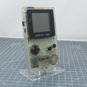 Könnte beinhalten: Transparente Game Boy Color Handheld-Spielkonsole. Die Konsole hat einen schwarzen Bildschirm und schwarze Tasten, mit dem Schriftzug "GAME BOY COLOR" auf der Vorderseite. Sie steht auf einem transparenten Acrylständer.