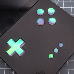 May include: A black handheld gaming device with a green and blue iridescent cross button, four round iridescent buttons, and three smaller iridescent round buttons.