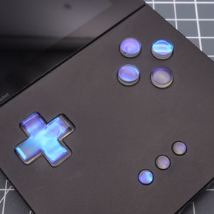 May include: A black handheld gaming device with a blue illuminated cross-shaped button and several round buttons. The device has a black screen and a gray background.