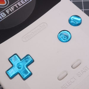 May include: A white handheld gaming console with blue buttons. The console has a directional pad, two buttons labelled "A" and "B", and two buttons labelled "Select" and "Start". The console also has the word "Nintendo" printed on it.