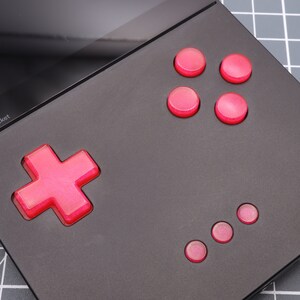 May include: A black handheld gaming device with a red cross-shaped button and six smaller red circular buttons. The device has a black screen.